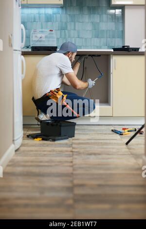 Les tuyaux doivent être entretenus. Jeune réparateur, plombier professionnel portant une ceinture à outils qui se croque sur le sol tout en fixant le tuyau de l'évier dans la cuisine Banque D'Images