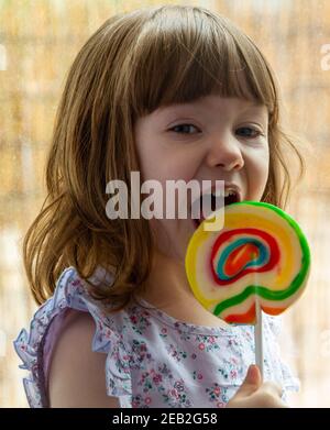 Portrait d'une jolie fille aux cheveux bruns et aux yeux bleus qui léchait une lollipop colorée par une fenêtre Banque D'Images