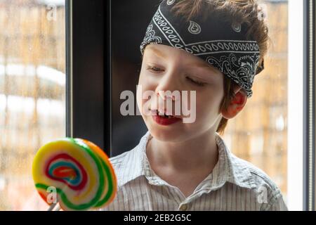 Portrait d'un adorable garçon aux cheveux rouges, aux yeux bleus, regardant un lollipop coloré près d'une fenêtre Banque D'Images