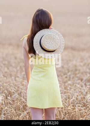 Jeune fille en robe jaune et avec chapeau de paille debout dans le champ de seigle d'orfroment. Concept d'été. Banque D'Images