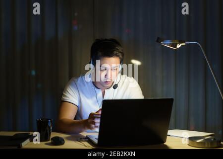 Les hommes asiatiques travaillant à la maison sont assis au travail avec leurs collègues ou leurs responsables par appel vidéo la nuit à la maison. Banque D'Images