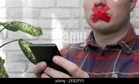 Un homme avec une bouche scellée avec un smartphone dans ses mains, un homme avec des lèvres scellées avec du ruban adhésif. Banque D'Images