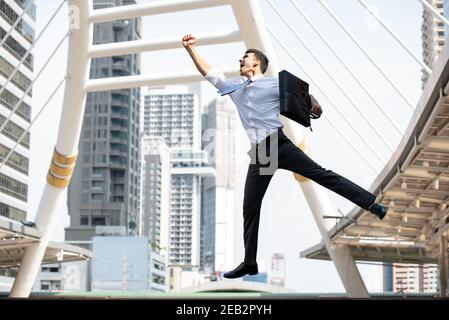 Jeune homme d'affaires asiatique heureux portant une mallette noire qui saute mi-air avec un bras relevé dans le geste de succès avec la ville bâtiment en arrière-plan Banque D'Images