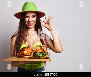 Jeune femme en jeans, corset et chapeau à large bord tient deux gros hamburger artisanal sur plateau en bois, gestes OK signe et nous entortiller sur fond gris. Rapide Banque D'Images