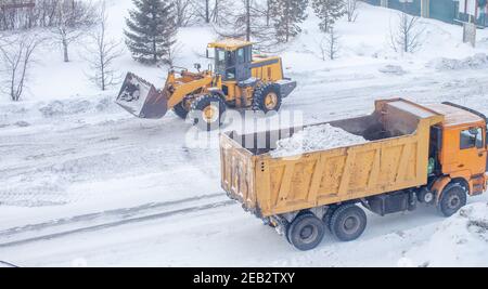 Le gros tracteur jaune nettoie la neige de la route et la charge dans le camion. Nettoyage et nettoyage des routes de la ville de la neige en hiver Banque D'Images