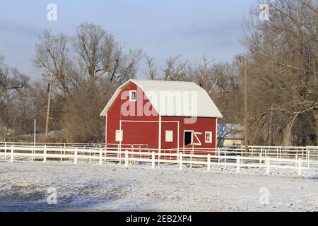 Grange rouge avec ciel bleu, arbres et clôture blanche au Kansas, lumineuse et colorée par une journée d'hiver. Banque D'Images