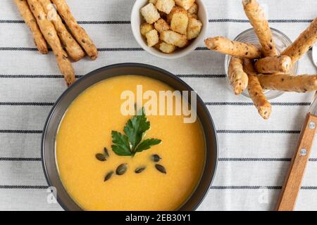 Un bol de soupe de potiron biologique crémeuse épicée maison sur fond de tissu blanc rayé avec des bâtonnets de pain, de la crème sure, du persil et des épices. Wi Banque D'Images