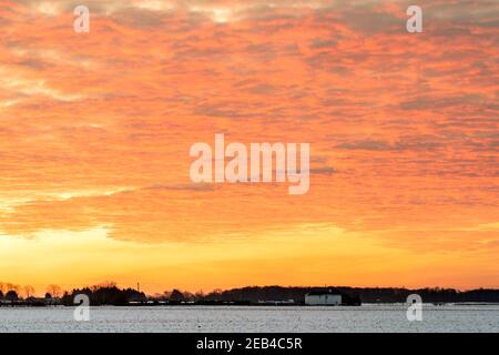 Le ciel de l'aube au-dessus d'un hedgerow lointain et des arbres à l'horizon un jour d'hiver après la chute de neige. L'horizon se trouve au bas du cadre. Le ciel est un jaune dramatique avec une couche de nuages rouges sous-éclairés, ce qui lui donne un ciel ondulé sur le feu look. Banque D'Images