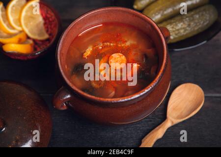 Soupe russe traditionnelle maison Solyanka avec viande, saucisses, légumes, cornichons et olives avec citron dans une casserole en céramique avec une cuillère en bois sur le woo sombre Banque D'Images