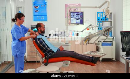 Infirmière mettant le bavoir dentaire à l'homme avant l'examen stomatologique en attente de stomatologiste médecin préparant le patient pour la chirurgie dentaire. Infirmière travaillant dans une clinique moderne d'orthodontie. Banque D'Images