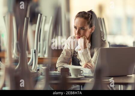 Une femme rateuse propriétaire de pub s'assoit au-dessus de l'ordinateur et du café en pensant à ce qu'il faut faire pendant que les affaires sont forcées de rester fermées en raison d'une pandémie. Banque D'Images