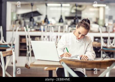 Une femme chef de restaurant s'assoit à la table après les heures d'ouverture de sa bibliothèque devant un ordinateur portable ouvert. Banque D'Images