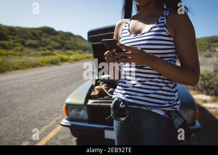 Femme de course mixte utilisant un smartphone debout sur la route à côté de la panne voiture avec capot ouvert Banque D'Images