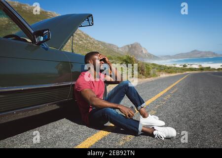 Homme afro-américain tenant un smartphone assis sur la route à côté de la panne voiture avec capot ouvert Banque D'Images