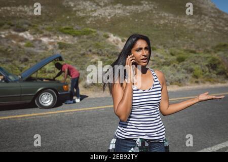 Femme de race mixte parlant avec un smartphone debout sur la route à côté voiture en panne avec capot ouvert Banque D'Images