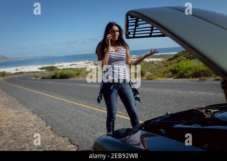 Femme de race mixte parlant avec un smartphone debout sur la route à côté voiture en panne avec capot ouvert Banque D'Images