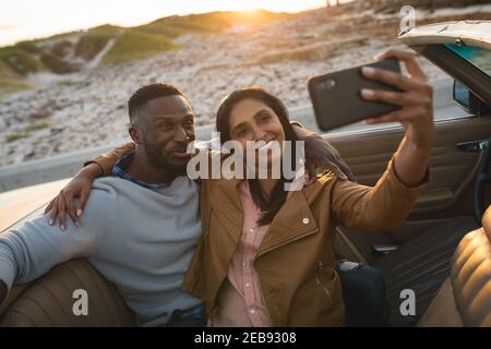 Divers couple assis dans une voiture convertible et de prendre un selfie Banque D'Images