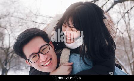 Un jeune homme donne un pigeyback à sa petite amie dans le parc le jour d'hiver enneigé. Un couple asiatique neuf s'est amusé dehors. Photo de haute qualité Banque D'Images