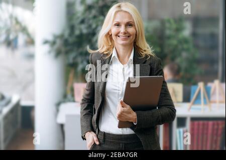 Portrait formel d'une femme d'affaires élégante et réussie, vêtue de costume, se tient dans un bureau moderne, tenant un ordinateur portable dans le bras. Charmante femme blonde mûre avec regarder et sourire directement à la caméra Banque D'Images