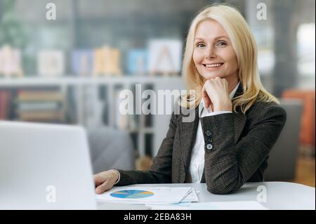 Portrait d'une femme d'affaires, d'un pdg ou d'un directeur d'âge moyen au bureau moderne. Élégante femme blonde confiante en costume élégant et habillé se trouve au bureau, regarde l'appareil photo et souriant sympathique Banque D'Images
