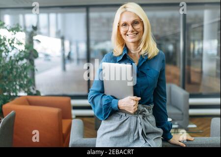 Portrait d'une femme d'affaires heureuse et confiante en eyaglasses et élégante tenue habillée. Femme d'âge moyen qui a réussi à regarder directement la caméra, souriant, tenant l'ordinateur portable dans les bras Banque D'Images