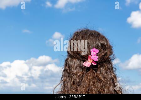 Une fille retournée avec des fleurs roses dans ses cheveux regarde dans le paysage contre un magnifique ciel bleu Banque D'Images