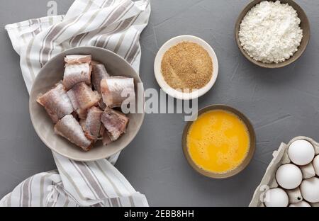 Dans un bol, des morceaux de merlu et des produits pour la cuisine frite, des œufs, de la farine et de la chapelure ont été présentés avec une serviette de cuisine sur fond gris Banque D'Images