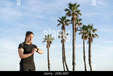 Fitness jeune femme regardant une montre intelligente après avoir courez avec belle palm Trees sur fond.technologie sportive et concept de style de vie sain avec co Banque D'Images