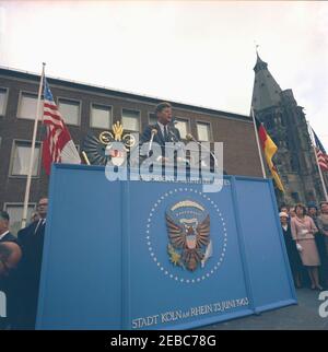 Voyage en Europe: Allemagne, Cologne: Ku00f6lner Rathaus (hôtel de ville), 10:55. Le président John F. Kennedy prononce une allocution devant Ku00f6lner Rathaus (hôtel de ville) à Cologne, Allemagne de l'Ouest (République fédérale); le directeur de la radio dans le secteur américain (RIAS) à Berlin et le traducteur du président Kennedy, Robert Lochner (masqué par un microphone), se tient à droite du président. Droite debout de la plate-forme speakersu2019: Eunice Kennedy Shriver; Cecilia McGhee, épouse de l'ambassadeur des États-Unis en Allemagne de l'Ouest, George McGhee. rn Banque D'Images