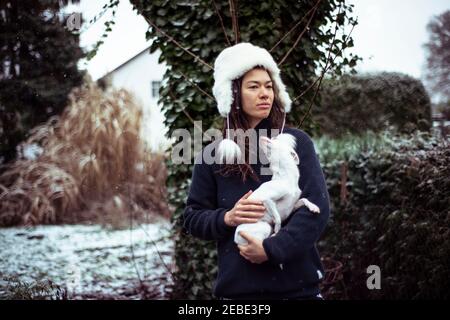 Personne dans le chapeau blanc molletonné tient dans la neige avec petit chien blanc dans la cour Banque D'Images