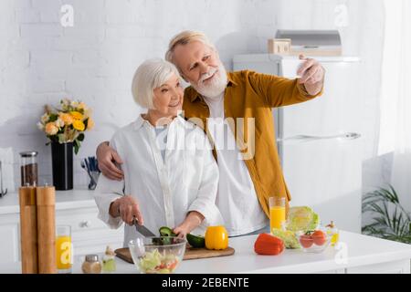 Homme senior qui prend le selfie sur son smartphone avec une femme souriante qui cuisine dans la cuisine Banque D'Images