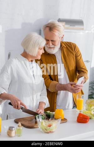 Homme senior tenant un smartphone près d'une femme souriante qui cuisine sur flou premier plan dans la cuisine Banque D'Images