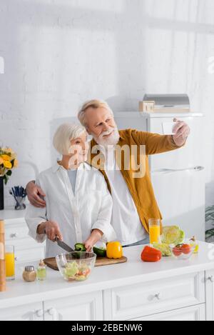 Couple de personnes âgées prenant le selfie tout en cuisant près de jus d'orange dedans cuisine Banque D'Images