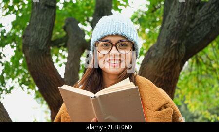 femme gaie en bonnet et lunettes souriant à l'appareil photo pendant tenue de livre dans le parc Banque D'Images