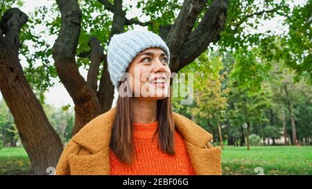 jeune femme en bonnet tricoté souriant tout en regardant loin dans stationnement Banque D'Images