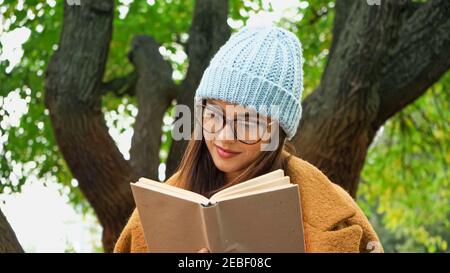 jeune femme en bonnet tricoté et lunettes de vue lisant le roman dans stationnement Banque D'Images