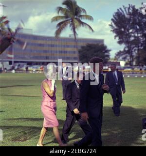 Discours devant la Brigade 2506 à Miami, 10:34AM. Le président John F. Kennedy et la première dame Jacqueline Kennedy traversent l'herbe après leur arrivée en hélicoptère (partiellement visible en haut à gauche) à Miami, en Floride, pour une cérémonie de présentation du 2506e drapeau de la brigade d'invasion cubaine au stade Orange Bowl. Marcher en premier plan (de gauche à droite): Mme Kennedy (portant des gants blancs et un foulard enveloppé sous son menton); le maire de Miami, Robert King High; le président Kennedy. Banque D'Images
