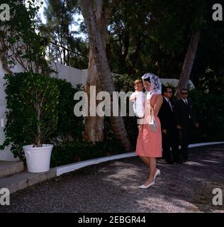 Président Kennedy u0026 famille assister à la messe à Palm Beach. La première dame Jacqueline Kennedy porte John F. Kennedy, Jr., alors qu'elle marche vers une chapelle privée à la résidence de l'ambassadeur Joseph P. Kennedy, Sr., à Palm Beach, Floride, pour le service du dimanche de Pâques. Les agents du service secret de la Maison Blanche, Ken Wiesman et Clint Hill, se tiennent en arrière-plan. Banque D'Images