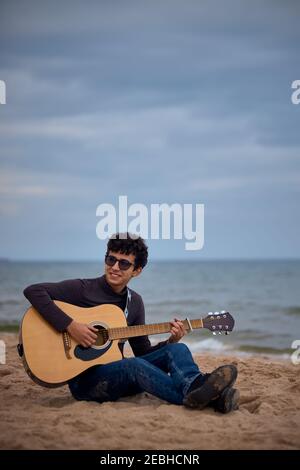 jeune adolescent caucasien jouant de la guitare acoustique sur la plage. lunettes de soleil et vêtements foncés. Banque D'Images