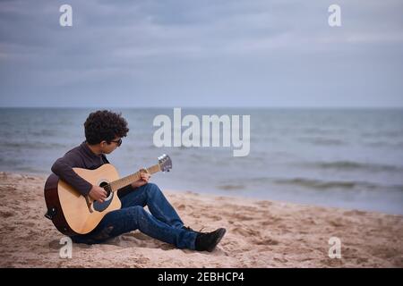 jeune adolescent caucasien jouant de la guitare acoustique sur la plage. lunettes de soleil et vêtements foncés. Banque D'Images