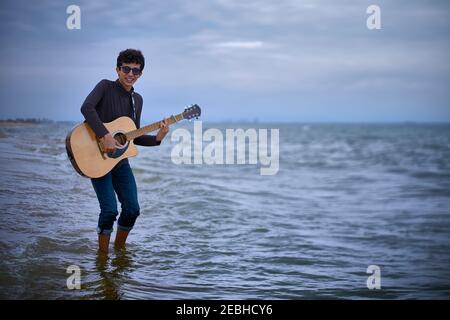 jeune adolescent caucasien jouant de la guitare acoustique sur la plage. lunettes de soleil et vêtements sombres. mouillant son pantalon avec une vague Banque D'Images