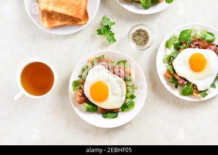 Des sandwichs ouverts avec des œufs frits, du bacon et des feuilles de légumes sur une assiette sur fond de pierre légère. Vue de dessus, plan d'appartement Banque D'Images