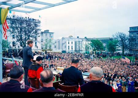 Voyage en Europe: Irlande, Galway: Président Kennedy à Eyre Square, remarques et départ, 11:55. Le président John F. Kennedy (à gauche, au pupitre) prononce un discours devant la foule rassemblée à Eyre Square à Galway, en Irlande, lors d'une cérémonie de bienvenue en son honneur ; le maire de Galway, Alderman Patrick D. Ryan (portant une robe rouge), siège à droite du président Kennedy ; d'autres sont non identifiés. A droite du pupitre (sur la table) se trouve le cercueil et le rouleau doré présentés au Président, dans le cadre de la cérémonie, lui accordant la liberté honoraire de la ville de Galway. Banque D'Images