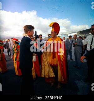 Voyage vers les États de l'Ouest : Honolulu, Hawaï, départ de l'aéroport international d'Honolulu, 5:35. Le président John F. Kennedy (à gauche, portant un lei fleuri) tremble les mains avec un homme non identifié (portant un costume traditionnel hawaïen rouge et jaune) avant de partir de l'aéroport international d'Honolulu à Honolulu, Hawaï ; trois femmes (portant des robes rouges et jaunes avec des chapeaux assortis) se tiennent à gauche. Banque D'Images