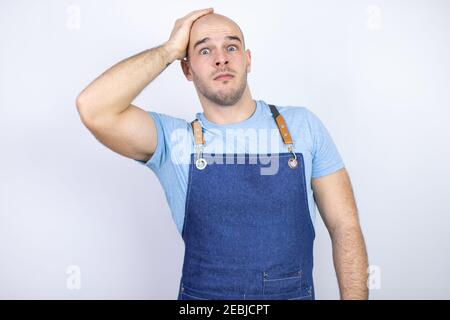 Jeune homme bald portant un tablier uniforme sur un fond blanc isolé mettre une main sur sa tête en souriant comme il l'avait fait quelque chose oublié Banque D'Images