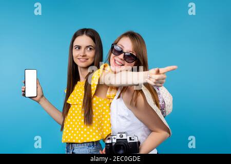 Deux jeunes femmes en vacances d'été sur fond bleu. Brunette tenant l'espace libre maquette de smartphone, pointant l'index dans la distance, blond Banque D'Images