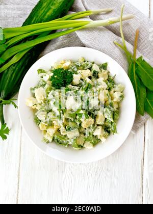 Salade de concombre, d'ostrel, de pommes de terre bouillies, d'œufs et d'herbes, garnie de mayonnaise dans une assiette blanche, de persil, d'oignons verts et de serviette en bois Banque D'Images