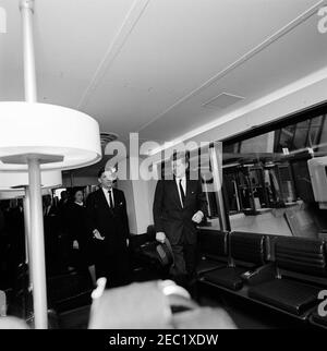 Cérémonies de dédicace, aéroport international de Dulles, 11:12. Le président John F. Kennedy (centre, chapeau de compagnie) traverse l'un des salons mobiles de l'aéroport international de Dulles au cours des cérémonies de dédicace de l'aéroport, nommé d'après le regretté secrétaire d'État John Foster Dulles. L'administrateur de l'Agence fédérale de l'aviation (FAA), Najeeb Halaby, et Aline B. Saarinen (veuve de l'architecte de l'aéroport, Eero Saarinen) suivent. Chantilly, Virginie. Banque D'Images