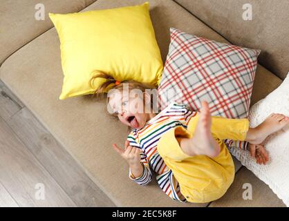 Une petite fille dans un pantalon jaune et un t-shirt coloré se trouve sur le dos de son canapé et montre sa langue, ce qui lui fait plaisir. Jeux pour enfants. C'est tout Banque D'Images