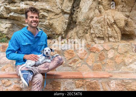 Jeune homme heureux attrayant épouse avec amour son mignon adopté mixte de Dalmatie chiot et assis sur le banc. Concept d'adoption des animaux de compagnie. Mur en pierre Banque D'Images